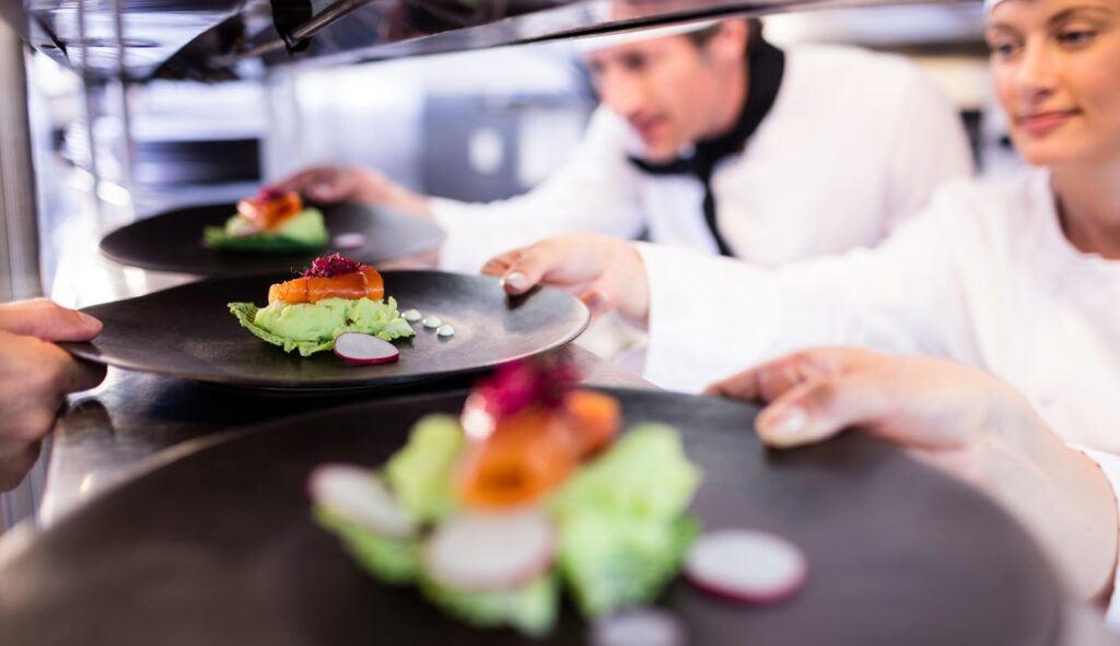 An image of two chefs handing over finished plates of food to a server. 